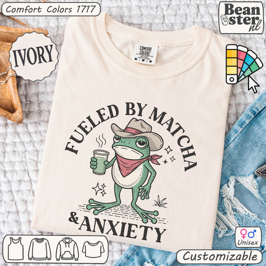 Beige t-shirt with a frog graphic and text 'Fueled by Matcha & Anxiety' on a textured surface.