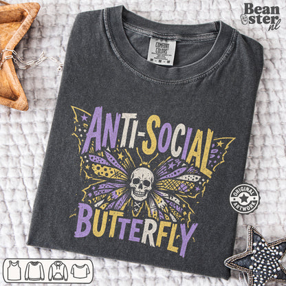 A dark gray heather t-shirt with a 'Anti-Social Butterfly' graphic, laid flat on a white textured blanket with decorative star-shaped props. The graphic features a purple and yellow butterfly with a skull in the center.