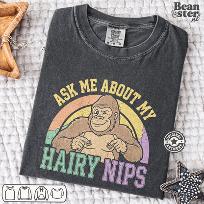 A dark charcoal gray graphic t-shirt laid flat on a textured white blanket. The shirt features a retro-style graphic of a smiling gorilla with a rainbow background and the text 'ASK ME ABOUT MY HAIRY NIPS' in yellow, green, and purple.