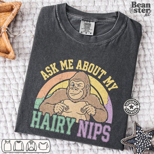 A dark charcoal gray graphic t-shirt laid flat on a textured white blanket. The shirt features a retro-style graphic of a smiling gorilla with a rainbow background and the text 'ASK ME ABOUT MY HAIRY NIPS' in yellow, green, and purple.