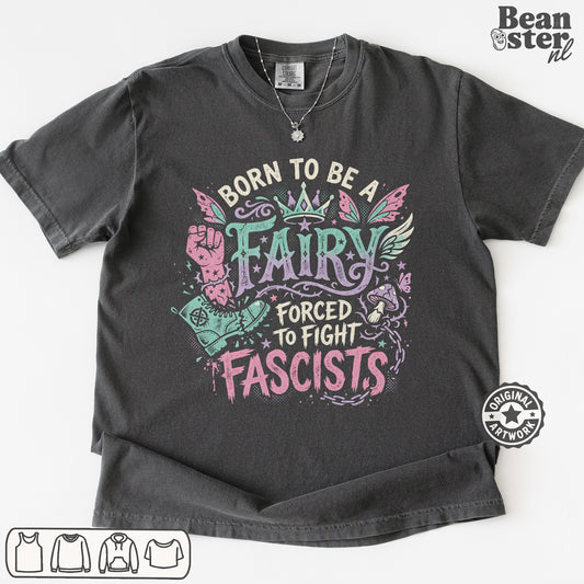 A dark charcoal gray graphic t-shirt laid flat on a white background. The shirt features a colorful graphic with the text 'BORN TO BE A FAIRY FORCED TO FIGHT FASCISTS' in pink, teal, and white.