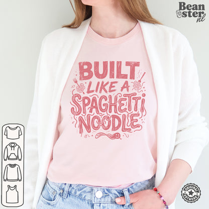 A woman wearing a light pink crewneck t-shirt with a red graphic that reads 'BUILT LIKE A SPAGHETTI NOODLE', styled with a white cardigan and blue jeans. The brand logo 'Bean ster nl' is visible in the top right corner.