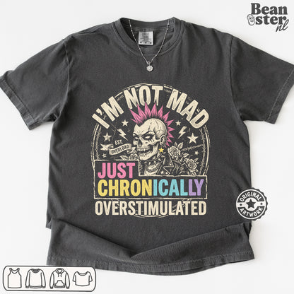 A dark gray graphic t-shirt featuring a skull with pink hair and the text 'I'M NOT MAD JUST CHRONICALLY OVERSTIMULATED'. 
