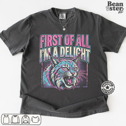 A dark gray graphic t-shirt featuring a stylized lynx cat illustration and the text 'FIRST OF ALL I'M A DELIGHT' in pink and teal. The t-shirt is displayed flat against a white background with a 'Beanster.