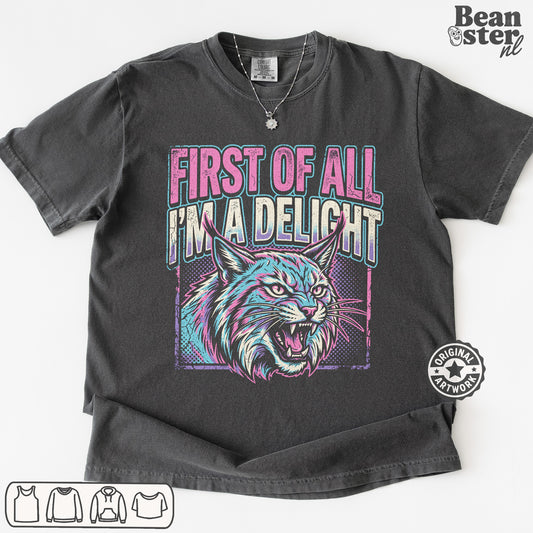 A dark gray graphic t-shirt featuring a stylized lynx cat illustration and the text 'FIRST OF ALL I'M A DELIGHT' in pink and teal. The t-shirt is displayed flat against a white background with a 'Beanster.