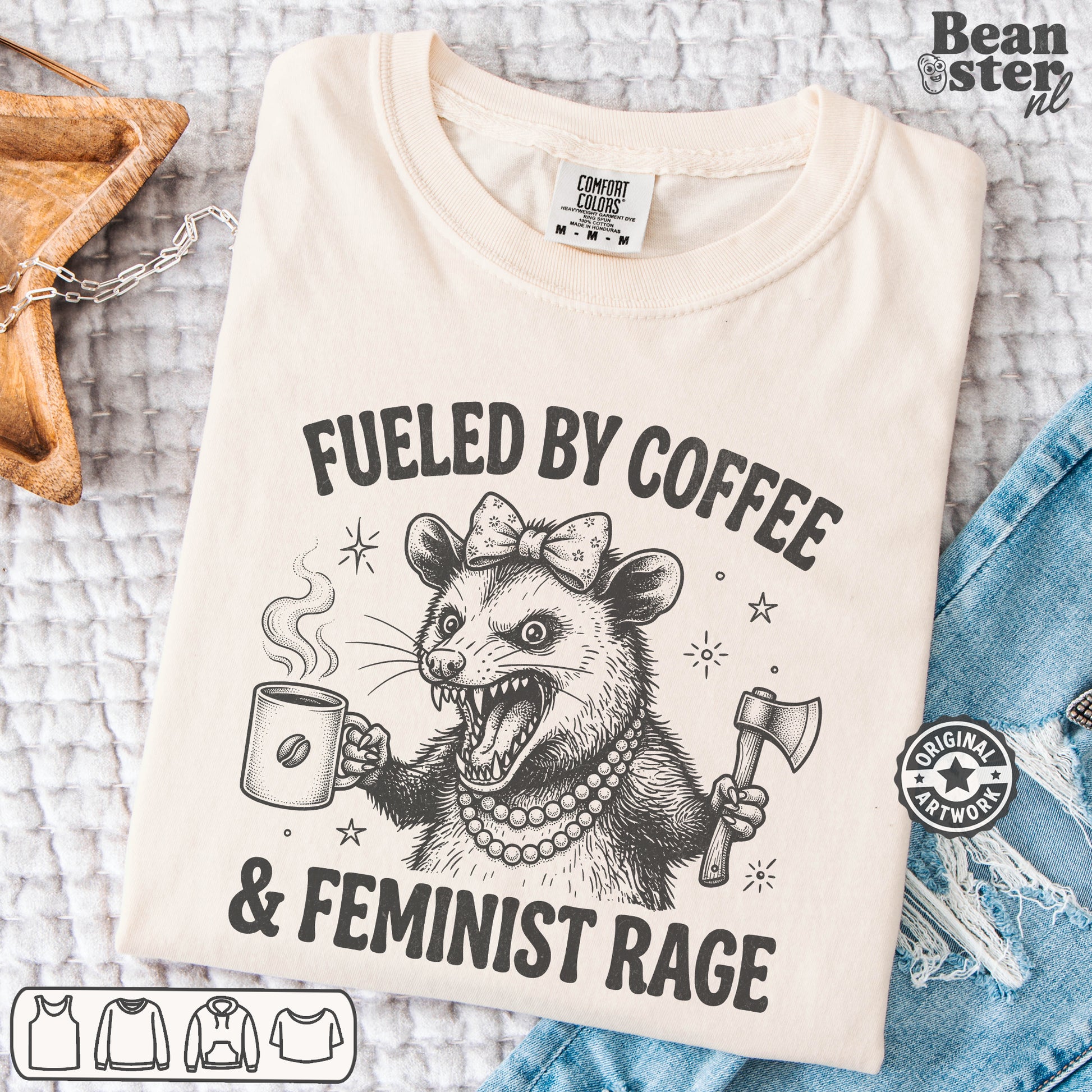 A folded cream-colored t-shirt with a black graphic print of a feminist opossum holding a coffee mug and an axe, with the text 'FUELED BY COFFEE & FEMINIST RAGE'. The shirt is styled in a flat-lay on a textured white blanket with a denim jacket, a woven bag, and a wooden spoon nearby.