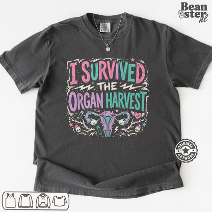 A dark gray graphic t-shirt featuring the text 'I SURVIVED THE ORGAN HARVEST' in pink, teal, and white, with a stylized uterus illustration below. 