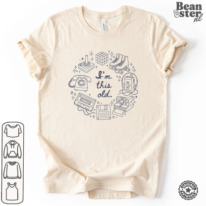 A light beige short-sleeve t-shirt with a dark blue graphic print of retro gaming items and the text 'I'm this old.' displayed on a white background.