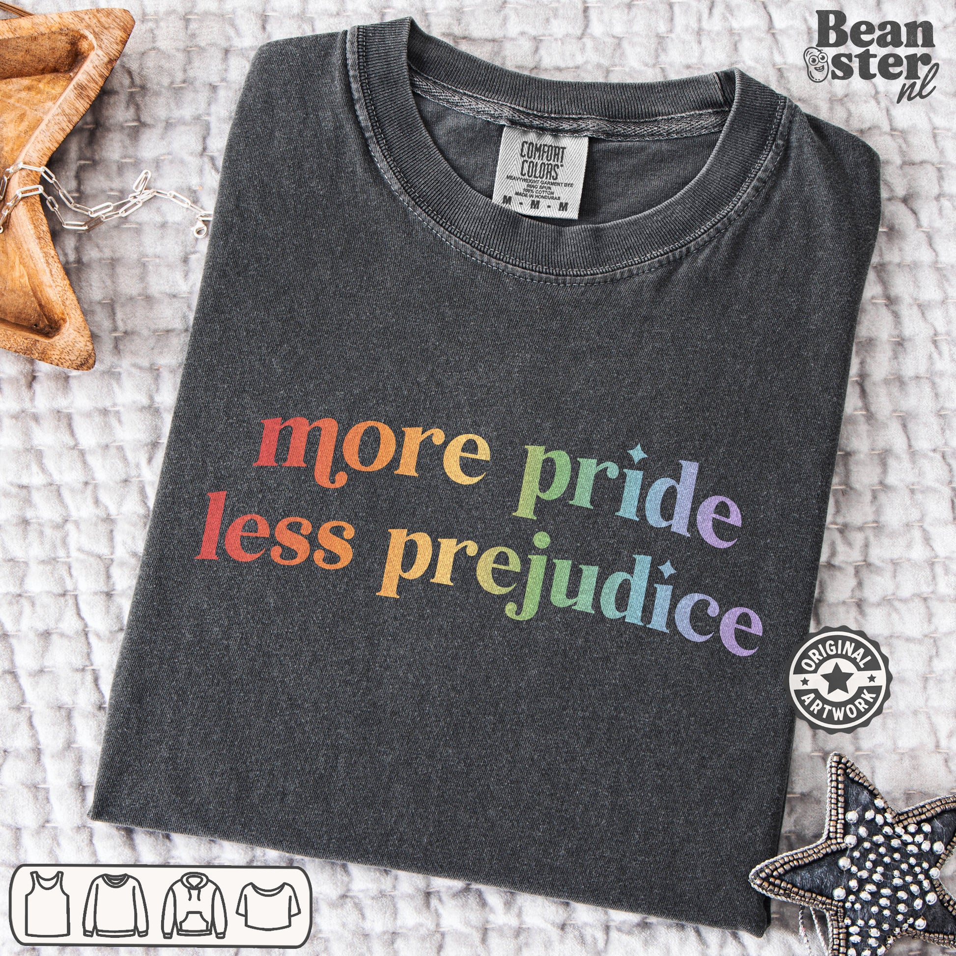 A dark charcoal gray t-shirt with the text 'more pride less prejudice' printed in a rainbow gradient, laid flat on a textured white blanket with decorative star-shaped props.