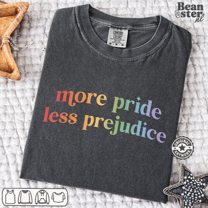 A dark charcoal gray t-shirt with the text 'more pride less prejudice' printed in a rainbow gradient, laid flat on a textured white blanket with decorative star-shaped props.