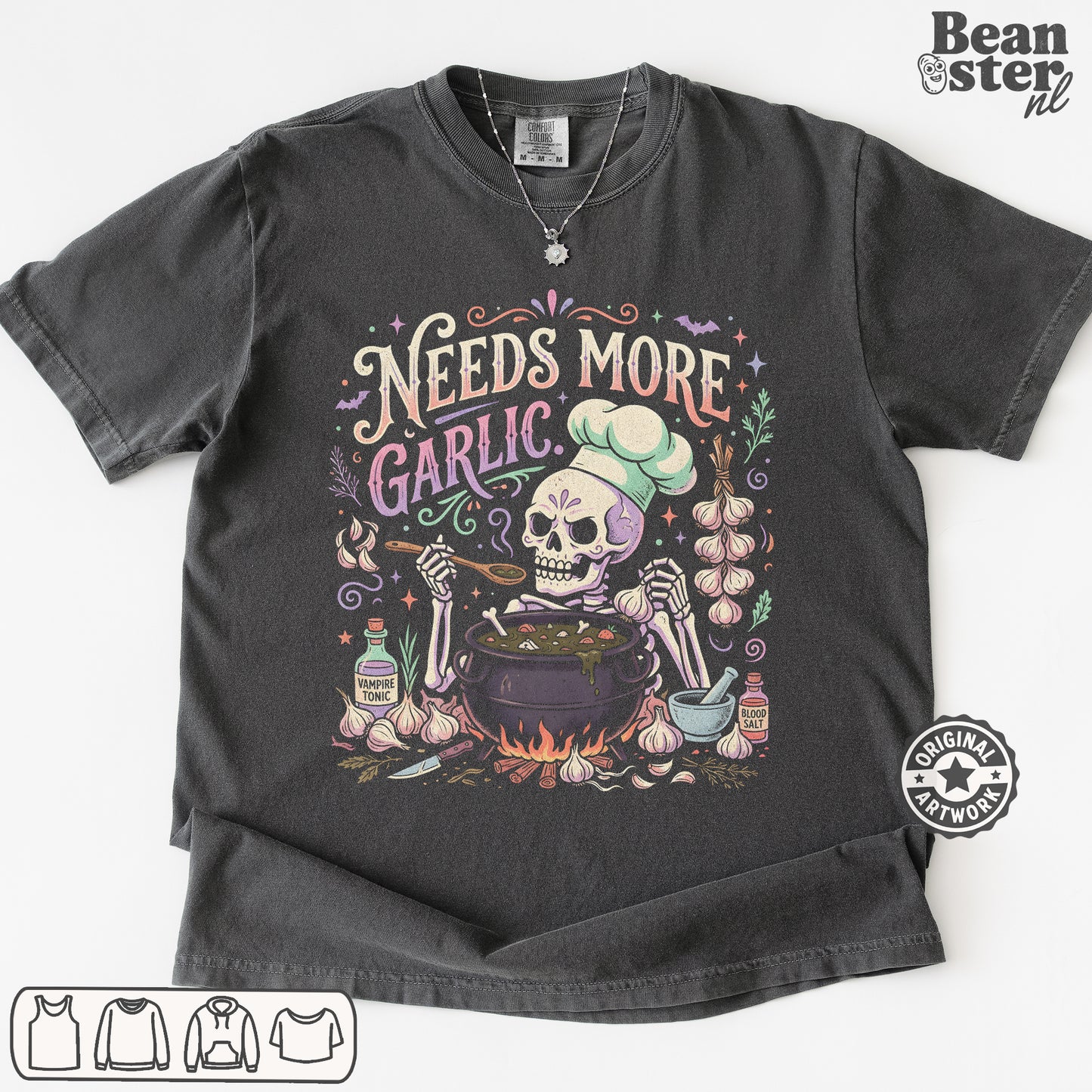 A dark charcoal gray graphic t-shirt featuring a Day of the Dead style illustration of a skeleton chef mixing ingredients in a pot, surrounded by garlic bulbs and the text 'Needs More Garlic'. The t-shirt is displayed flat against a white background with a 'Beanster.