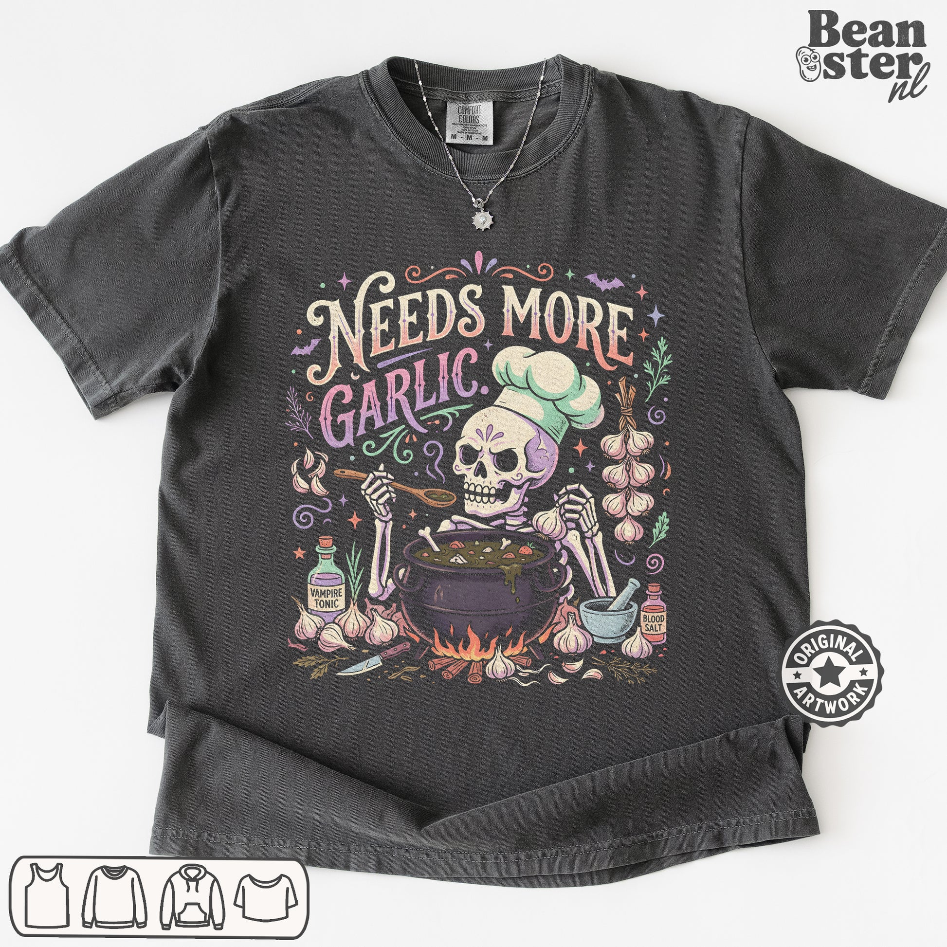 A dark charcoal gray graphic t-shirt featuring a Day of the Dead style illustration of a skeleton chef mixing ingredients in a pot, surrounded by garlic bulbs and the text 'Needs More Garlic'. The t-shirt is displayed flat against a white background with a 'Beanster.