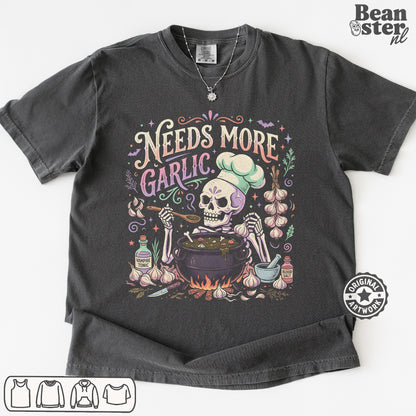 A dark charcoal gray graphic t-shirt featuring a Day of the Dead style illustration of a skeleton chef mixing ingredients in a pot, surrounded by garlic bulbs and the text 'Needs More Garlic'. The t-shirt is displayed flat against a white background with a 'Beanster.
