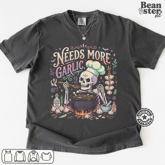 A dark charcoal gray graphic t-shirt featuring a Day of the Dead style illustration of a skeleton chef mixing ingredients in a pot, surrounded by garlic bulbs and the text 'Needs More Garlic'. The t-shirt is displayed flat against a white background with a 'Beanster.