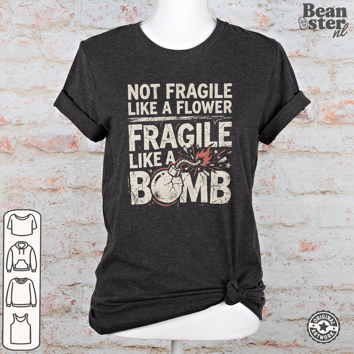 A dark charcoal gray graphic t-shirt displayed on a white mannequin torso against a faux white brick wall background. The shirt features a distressed white text print that reads 'NOT FRAGILE LIKE A FLOWER FRAGILE LIKE A BOMB' with a graphic of a cracked bomb.