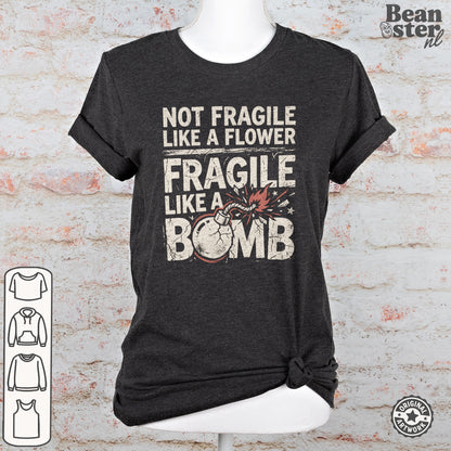 A dark charcoal gray graphic t-shirt displayed on a white mannequin torso against a faux white brick wall background. The shirt features a distressed white text print that reads 'NOT FRAGILE LIKE A FLOWER FRAGILE LIKE A BOMB' with a graphic of a cracked bomb.