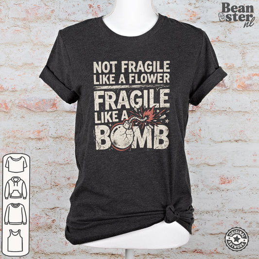 A dark charcoal gray graphic t-shirt displayed on a white mannequin torso against a faux white brick wall background. The shirt features a distressed white text print that reads 'NOT FRAGILE LIKE A FLOWER FRAGILE LIKE A BOMB' with a graphic of a cracked bomb.