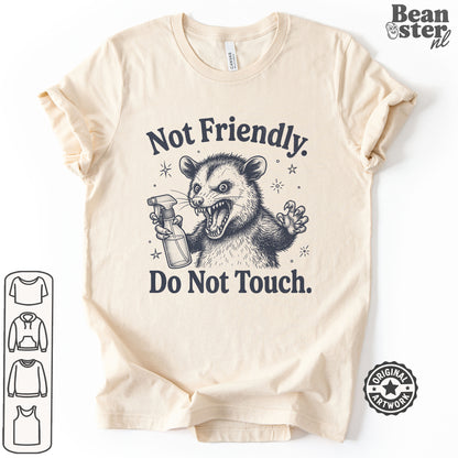 A beige short-sleeve t-shirt with a dark graphic of a mischievous possum holding a spray bottle, accompanied by the text 'Not Friendly. Do Not Touch.