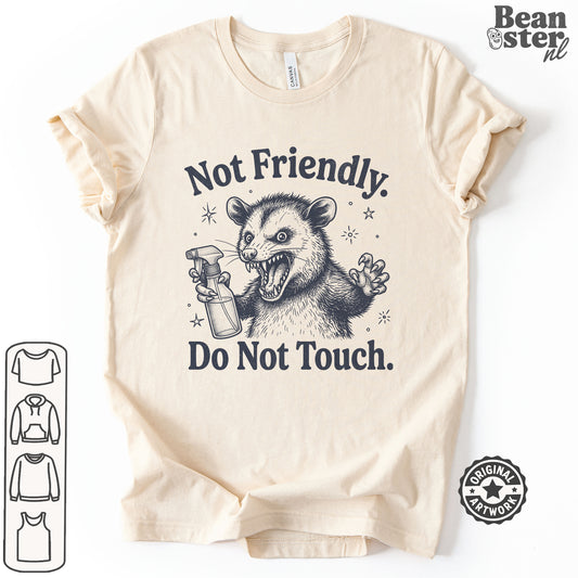 A beige short-sleeve t-shirt with a dark graphic of a mischievous possum holding a spray bottle, accompanied by the text 'Not Friendly. Do Not Touch.
