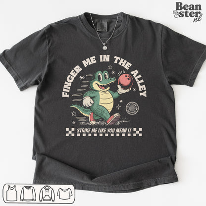 Black t-shirt with a graphic of a crocodile holding a bowling ball and text 'Finger Me in the Alley, Strike Me Like You Mean It' on a white background.