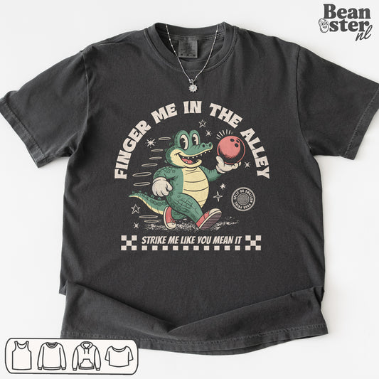 Black t-shirt with a graphic of a crocodile holding a bowling ball and text 'Finger Me in the Alley, Strike Me Like You Mean It' on a white background.
