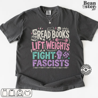 A dark gray graphic t-shirt laid flat on a white background. The shirt features a colorful distressed print with the text 'READ BOOKS LIFT WEIGHS FIGHT FASCISTS' accompanied by illustrations of books, skulls, a dumbbell, a raised fist, and a swastika symbol.