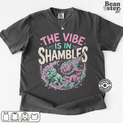 A dark charcoal gray graphic t-shirt laid flat on a white background. The shirt features a colorful print with the text 'THE VIBE IS IN SHAMBLFS' in pink, teal, and white, above an illustration of a purple and teal shambler dinosaur.