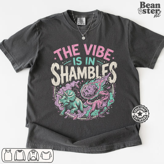 A dark charcoal gray graphic t-shirt laid flat on a white background. The shirt features a colorful print with the text 'THE VIBE IS IN SHAMBLFS' in pink, teal, and white, above an illustration of a purple and teal shambler dinosaur.