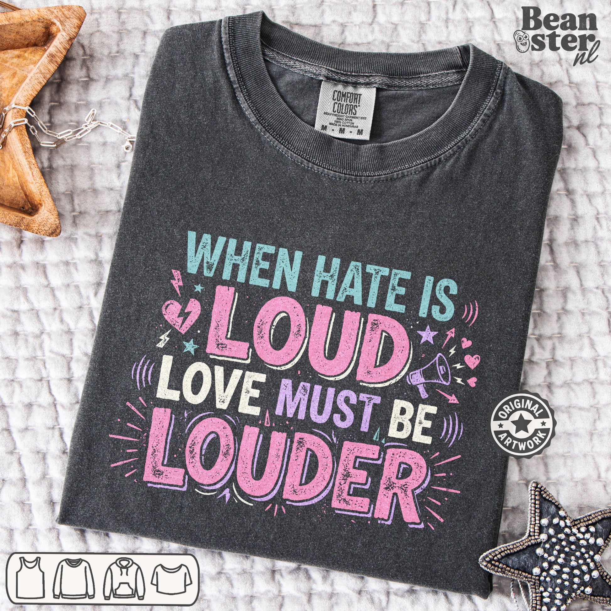 A dark charcoal gray graphic t-shirt is laid flat on a textured white blanket. The shirt features a colorful distressed text design that reads 'WHEN HATE IS LOUD LOVE MUST BE LOUDER' in shades of pink, light blue, and white.