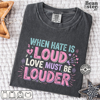 A dark charcoal gray graphic t-shirt is laid flat on a textured white blanket. The shirt features a colorful distressed text design that reads 'WHEN HATE IS LOUD LOVE MUST BE LOUDER' in shades of pink, light blue, and white.