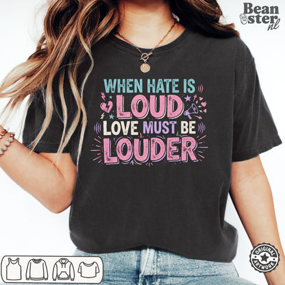 A woman wearing a black graphic t-shirt with the text 'WHEN HATE IS LOUD LOVE MUST BE LOUDER' in pink, purple, and teal lettering. The photo is cropped from the chest down, showing the fit of the shirt, with a 'Beanster.